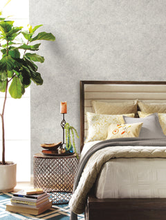 A cozy bedroom featuring York Wallcoverings’ Floret Wallpaper, a bed adorned with decorative pillows, a side table displaying a candle, plant, and books, and a large potted plant by the window. The space showcases a neutral color palette complemented by a geometric rug.