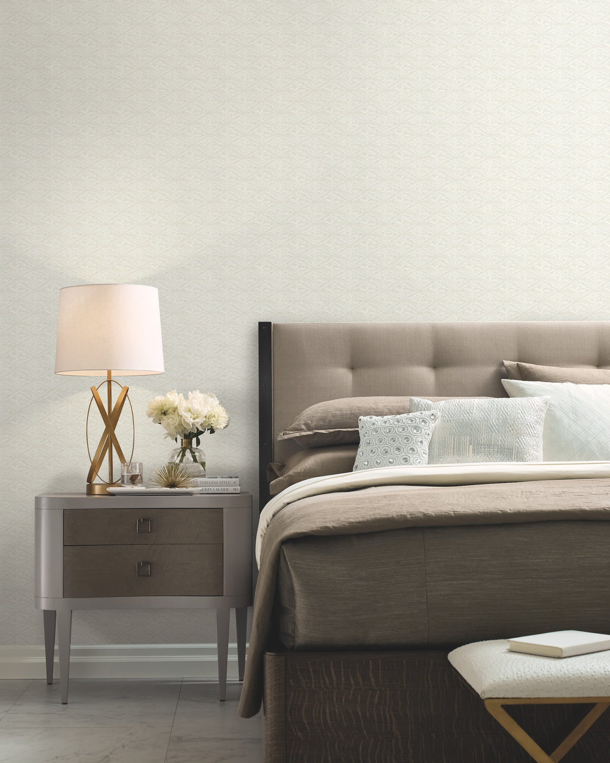 A modern bedroom featuring a tufted headboard, neutral bedding, and a gray nightstand holding a lamp, white flowers, and a book. At the foot of the bed sits a white textured bench. The walls are adorned with York Wallcoverings’ High Society Wallpaper in a light patterned design.