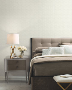A modern bedroom featuring a tufted headboard, neutral bedding, and a gray nightstand holding a lamp, white flowers, and a book. At the foot of the bed sits a white textured bench. The walls are adorned with York Wallcoverings’ High Society Wallpaper in a light patterned design.