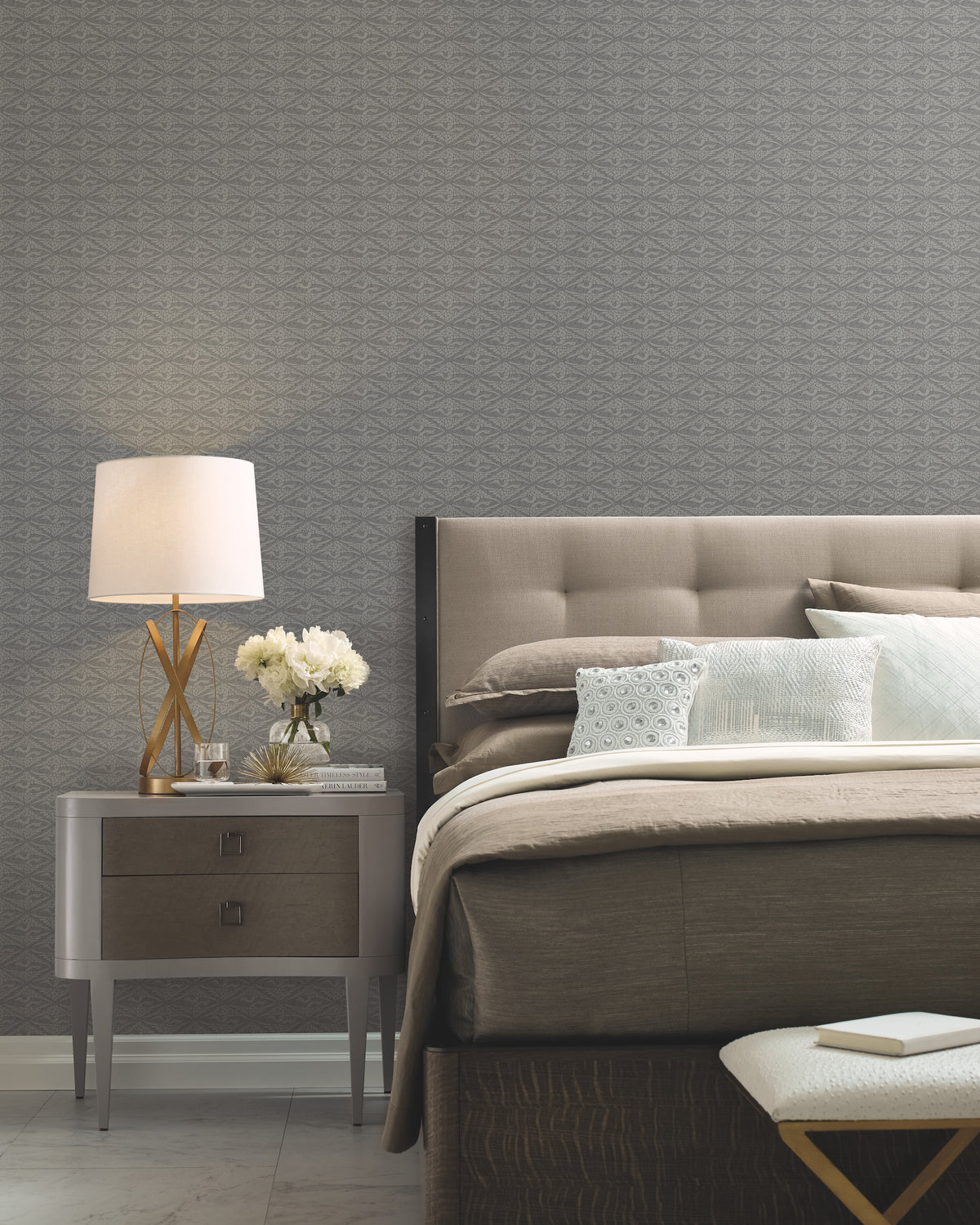 A modern bedroom featuring a gray padded headboard, neatly arranged pillows, a bedside table with a lamp, white flowers, and books, plus a patterned accent wall adorned with York Wallcoverings’ High Society Wallpaper.