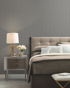 A modern bedroom featuring a gray padded headboard, neatly arranged pillows, a bedside table with a lamp, white flowers, and books, plus a patterned accent wall adorned with York Wallcoverings’ High Society Wallpaper.