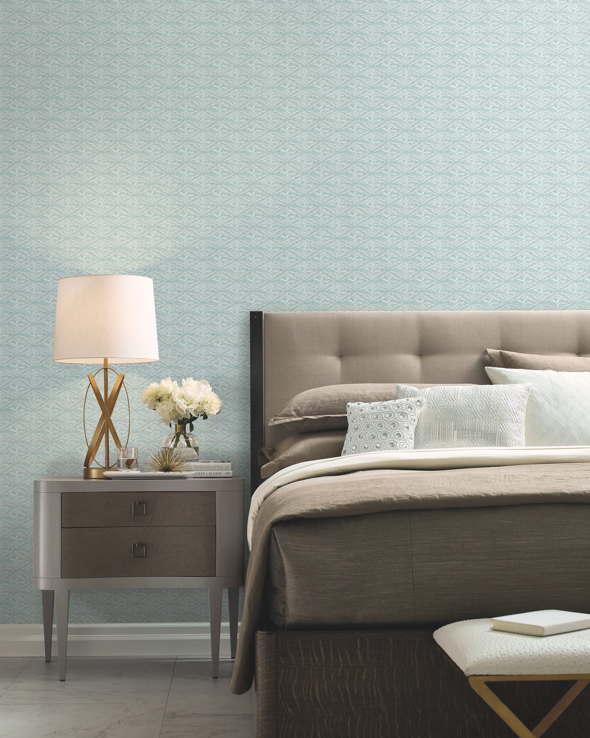 Modern bedroom with a tufted headboard, neutral bedding, and a nightstand topped with a lamp, books, and white flowers—all set against York Wallcoverings’ High Society Wallpaper in light blue.