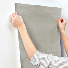 Hands applying a sheet of grey, geometric patterned In Clay Wallpaper by York Wallcoverings to a white wall.