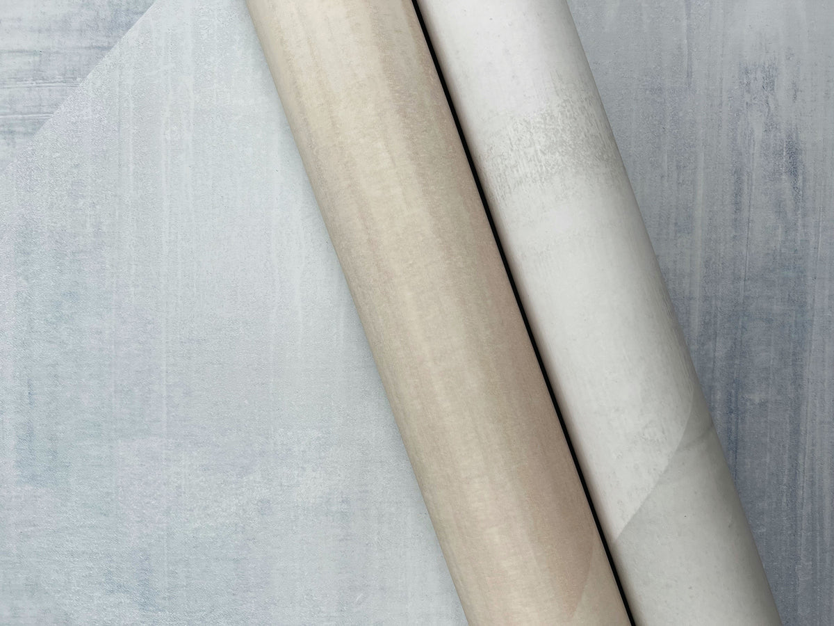 Two rolls of In Clay Wallpaper with a textured, mottled appearance resting on a piece of the same wallpaper.