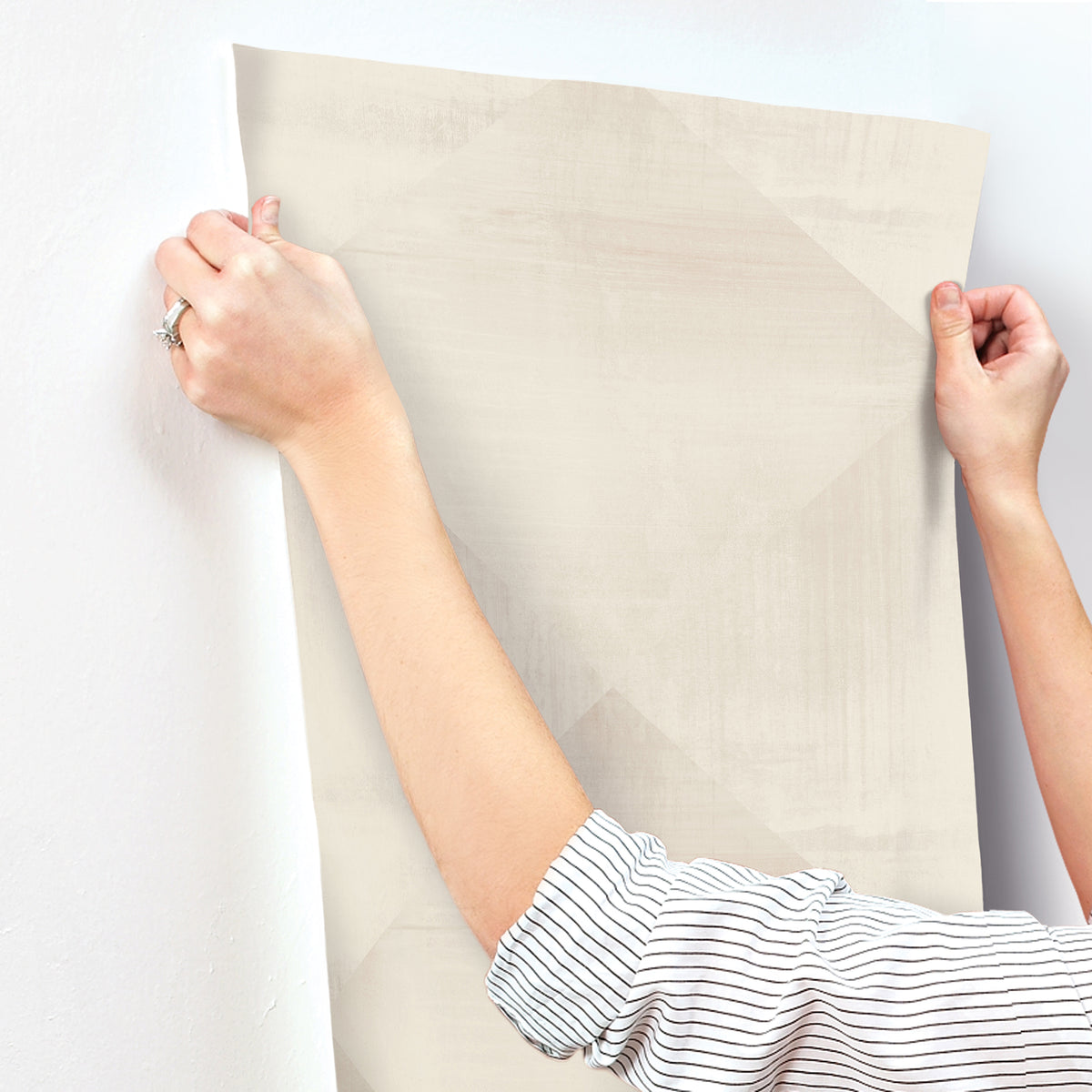 Hands apply a sheet of beige In Clay Wallpaper with a subtle geometric pattern to a white wall.