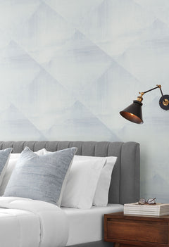 In Clay Wallpaper in light blue and white with a soft, brushed geometric diamond pattern behind a bed with a gray upholstered headboard.