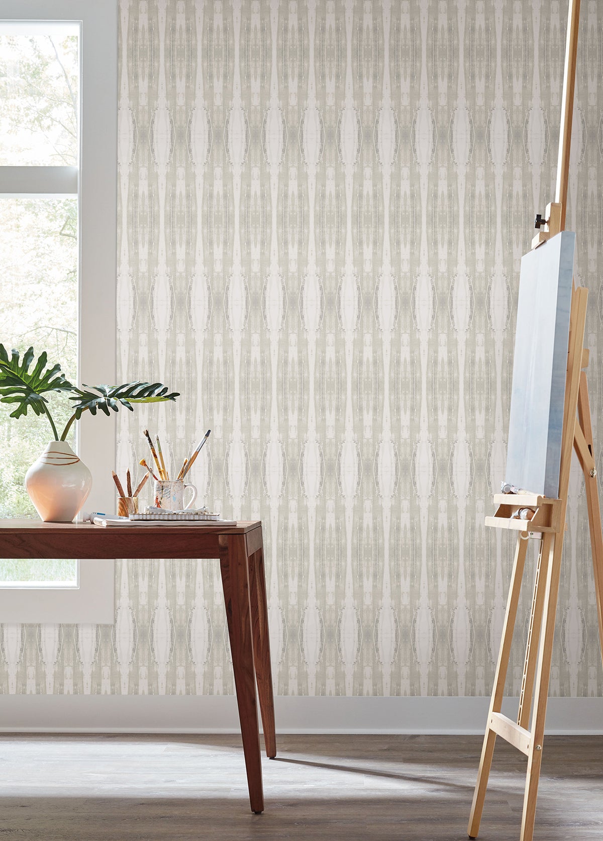 A wooden table adorned with art supplies and a plant sits by a window; an empty easel stands nearby against a backdrop of York Wallcoverings’ Escalante Wallpaper.
