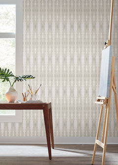 A wooden table adorned with art supplies and a plant sits by a window; an empty easel stands nearby against a backdrop of York Wallcoverings’ Escalante Wallpaper.