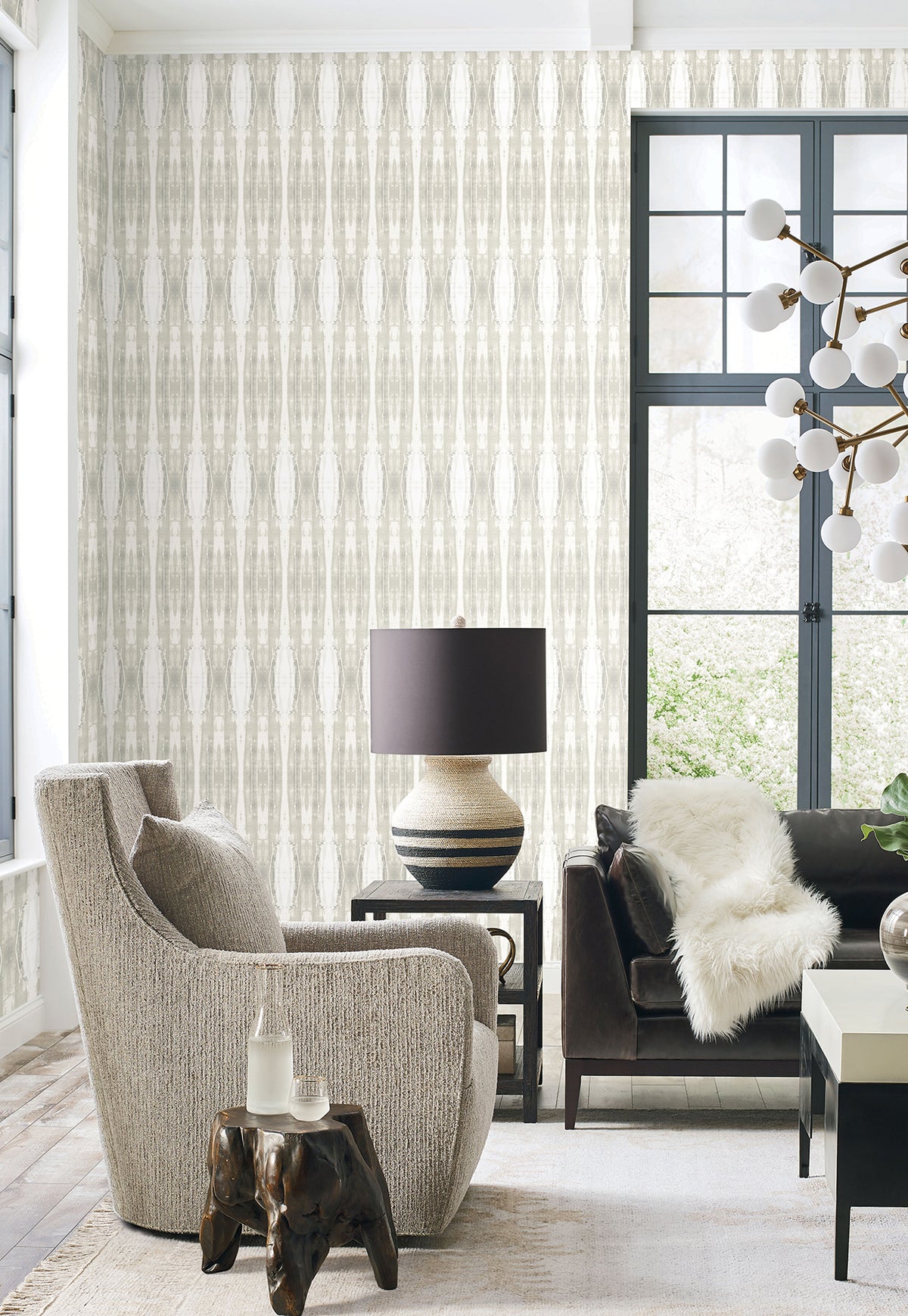 Modern living room with a gray armchair, dark sofa with white fur throw, round lamp on a side table, unique chandelier, wooden side table, large windows, and York Wallcoverings’ Escalante Wallpaper.