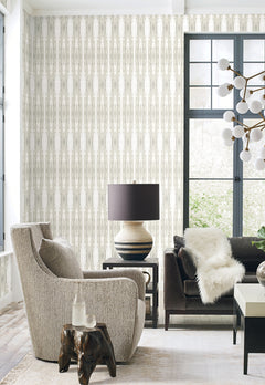 Modern living room with a gray armchair, dark sofa with white fur throw, round lamp on a side table, unique chandelier, wooden side table, large windows, and York Wallcoverings’ Escalante Wallpaper.