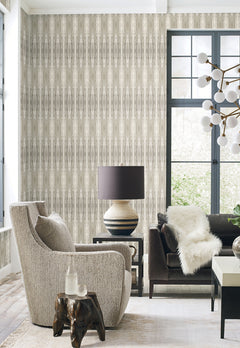 A modern living room featuring a textured gray chair, dark sofa with a white fur throw, Escalante Wallpaper by York Wallcoverings, a unique lamp on a small table, a window with black trim, and a white branched chandelier overhead.