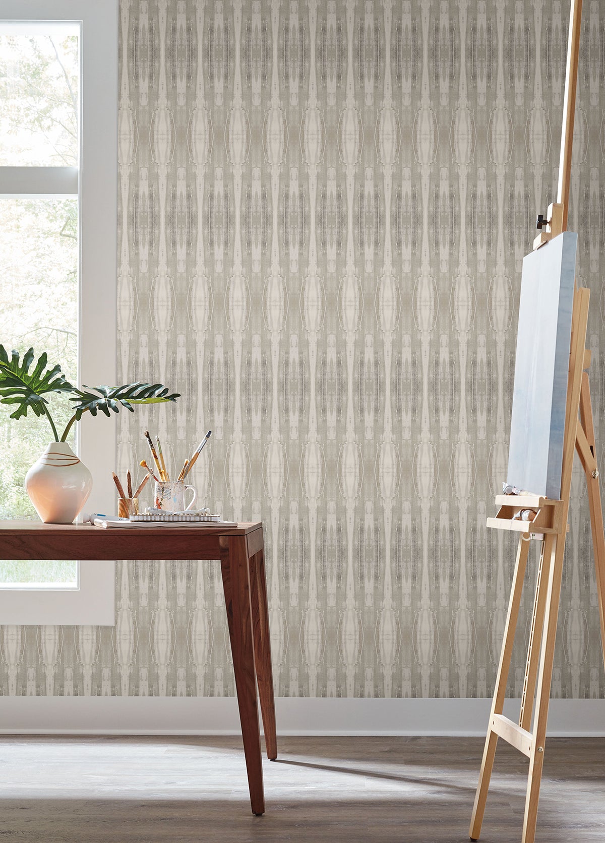 A wooden desk with art supplies and a plant sits by a window; an easel stands nearby against Escalante Wallpaper by York Wallcoverings in a bright room.
