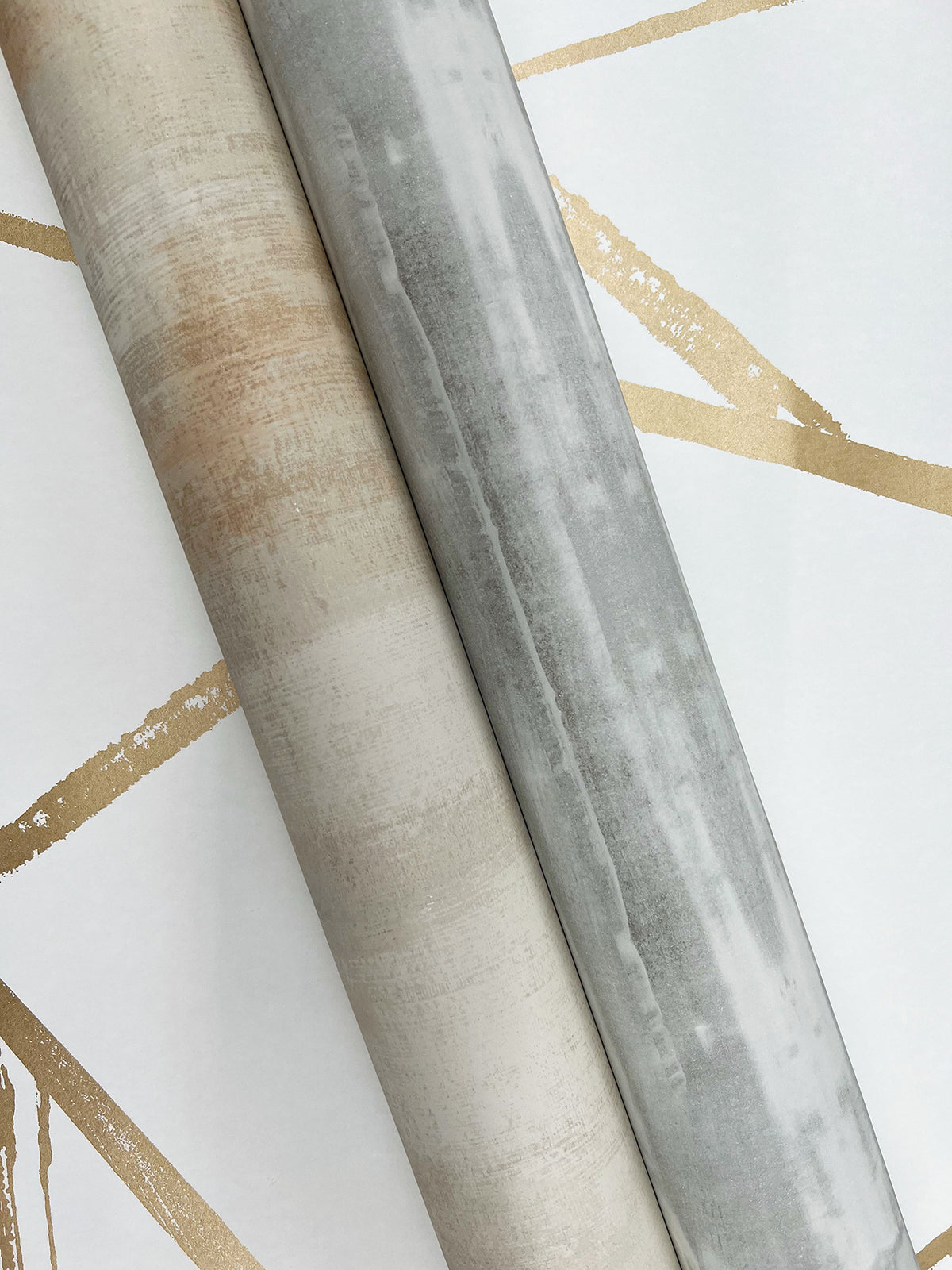 Two rolls of Escalante Wallpaper, one tan and one gray, resting on a white surface featuring a geometric pattern of gold lines.
