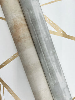 Two rolls of Escalante Wallpaper, one tan and one gray, resting on a white surface featuring a geometric pattern of gold lines.