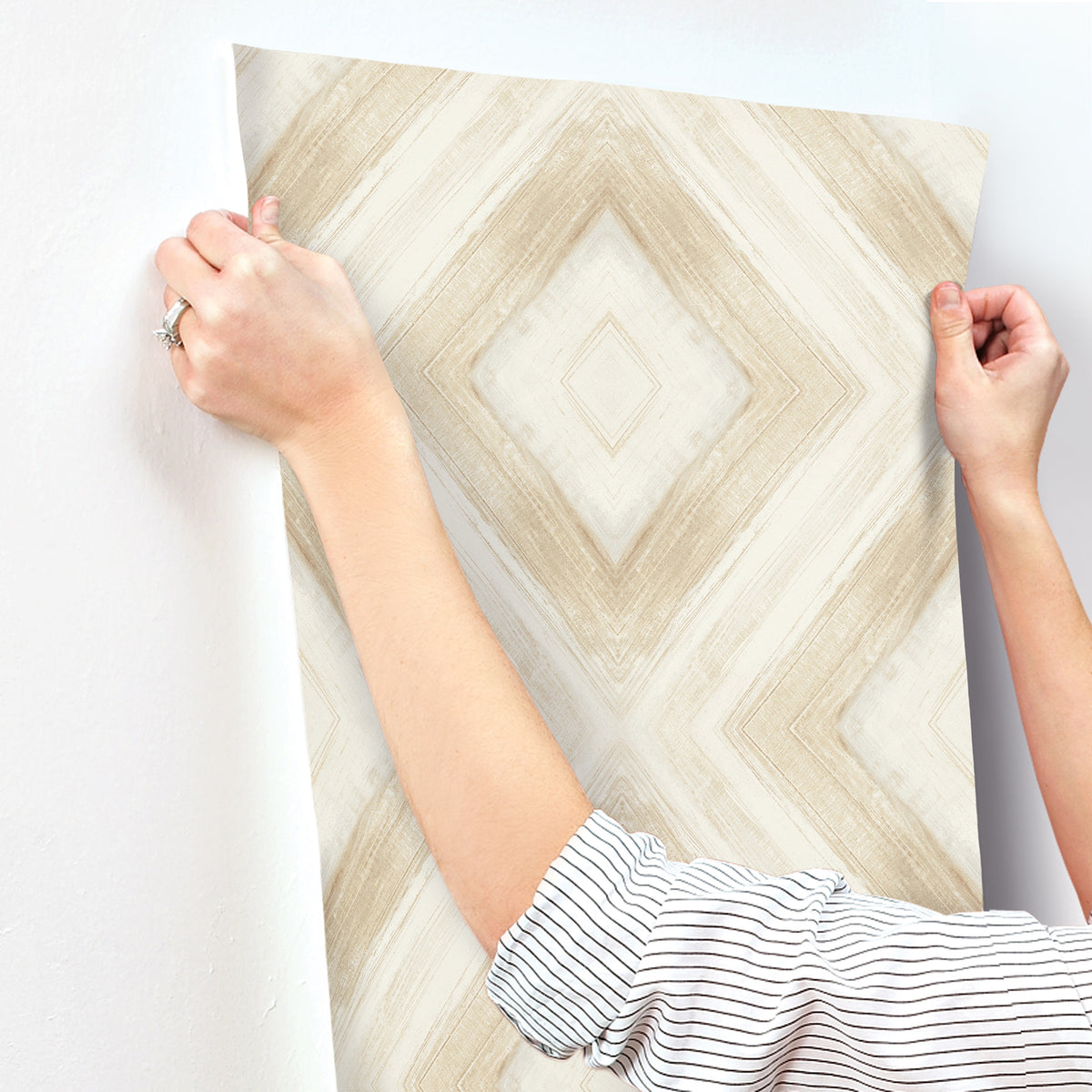 Hands applying a sheet of Ballad Wallpaper with a beige and white diamond-patterned brushstroke design to a white wall.