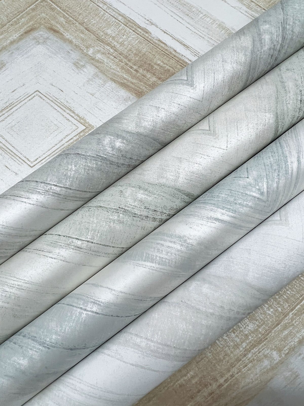 Three rolls of metallic silver swirling Ballad Wallpaper resting on a sheet of wallpaper featuring a geometric diamond design.