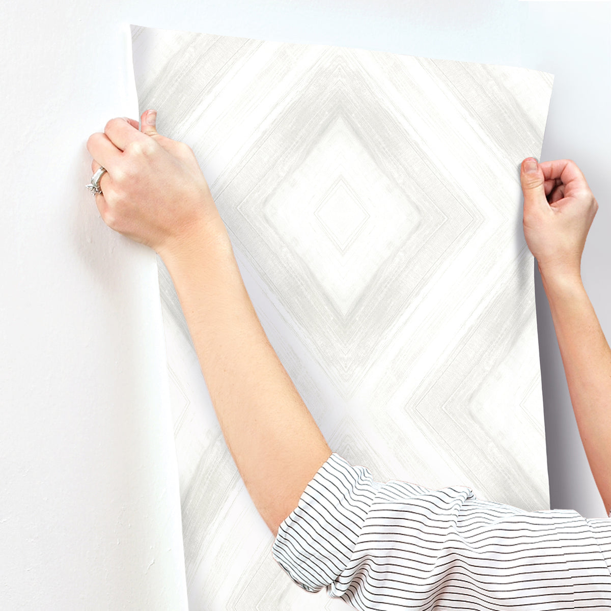 Hands applying a sheet of off-white Ballad Wallpaper with a subtle grey diamond pattern to a wall.