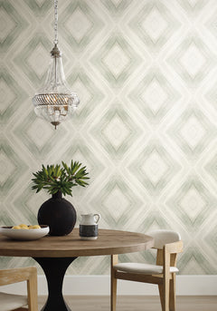 Ballad Wallpaper featuring a repeating sage green diamond pattern behind a round wooden table, a dark vase with greenery, and a glass pendant light.