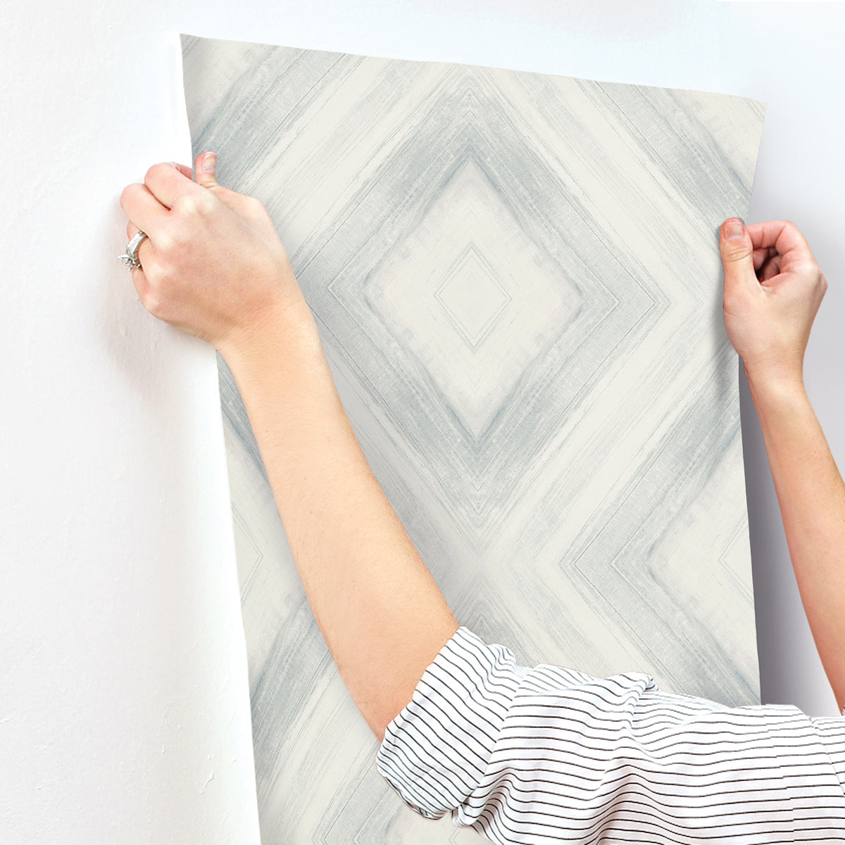 Hands holding a strip of grey and white geometric diamond Ballad Wallpaper against a wall.