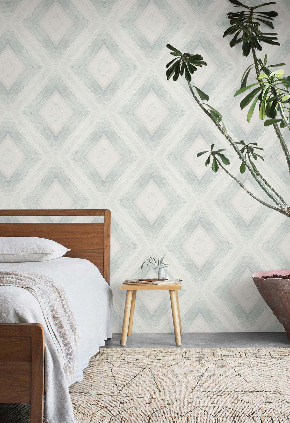 Ballad Wallpaper in a repeating blue and white diamond pattern behind a wooden bed and a side table with a potted plant.