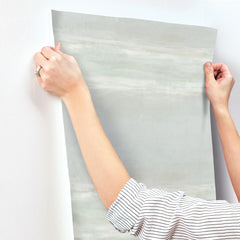 Hands applying a sheet of pale green, brush-stroked Serene Reflection Wallpaper by York Wallcoverings to a white wall.