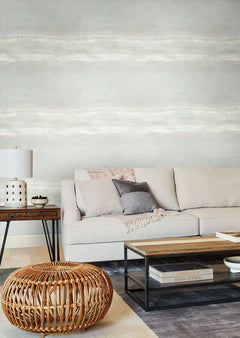 A living room with a beige sofa, a low wooden coffee table, and a rattan ottoman against a wall covered in Serene Reflection Wallpaper by York Wallcoverings, featuring a light, horizontal-brushed texture.