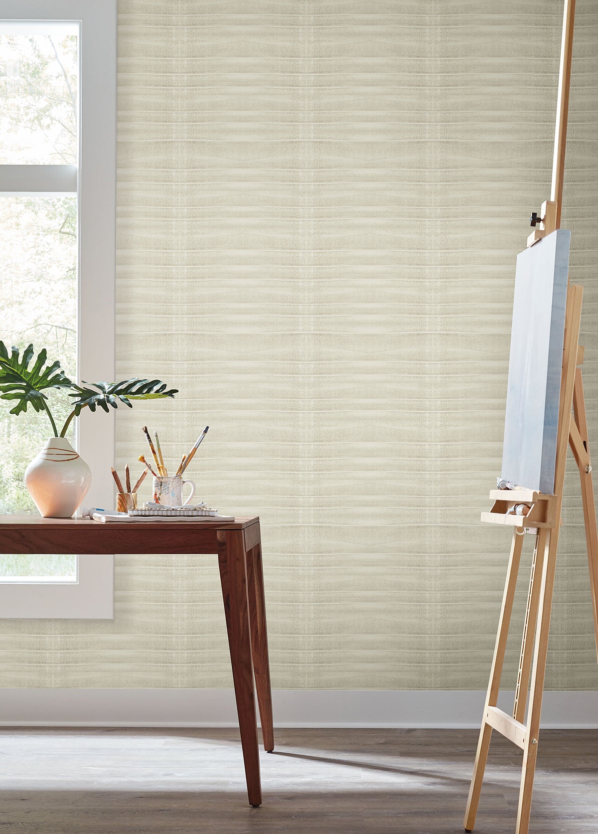 Grey Stone Wallpaper in light beige with a horizontal, subtle wave texture installed on a wall behind an artist’s easel and a side table.