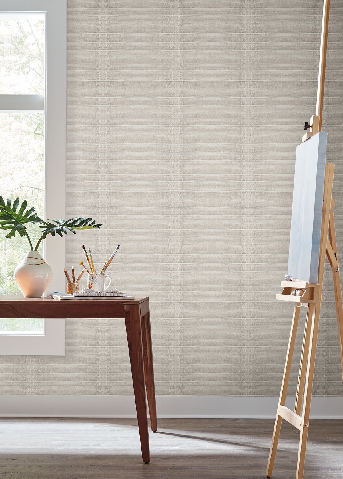 Grey Stone Wallpaper with a subtle light grey horizontal ribbed pattern covering a wall in a room featuring a wooden table, a plant, and an artist’s easel.