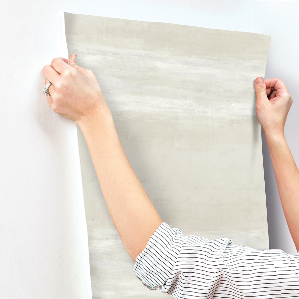 Hands holding a sheet of light grey, textured Grey Stone Wallpaper against a white wall.