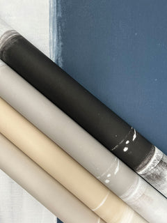 Four rolls of Ink Wash Wallpaper by York Wallcoverings displayed diagonally against a backdrop of deep blue and white panels, showing gradients of charcoal, grey, beige, and light cream.