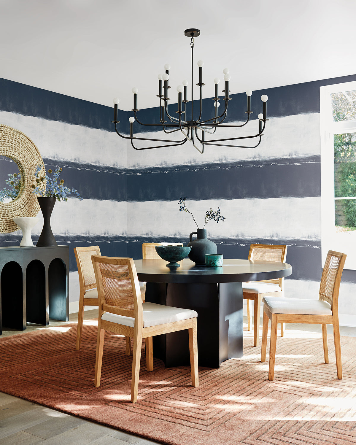 A dining room with a round black table and four cane-back chairs, featuring walls covered in York Wallcoverings Ink Wash Wallpaper with a dark navy and white horizontal design.