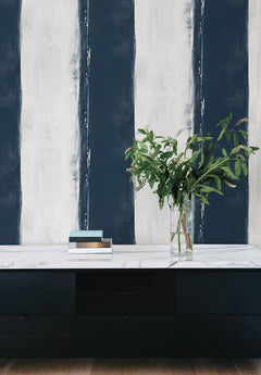 Ink Wash Wallpaper features wide, vertical navy blue and white stripes with a distressed, painted texture behind a black cabinet with a marble top, books, and a glass vase of greenery.