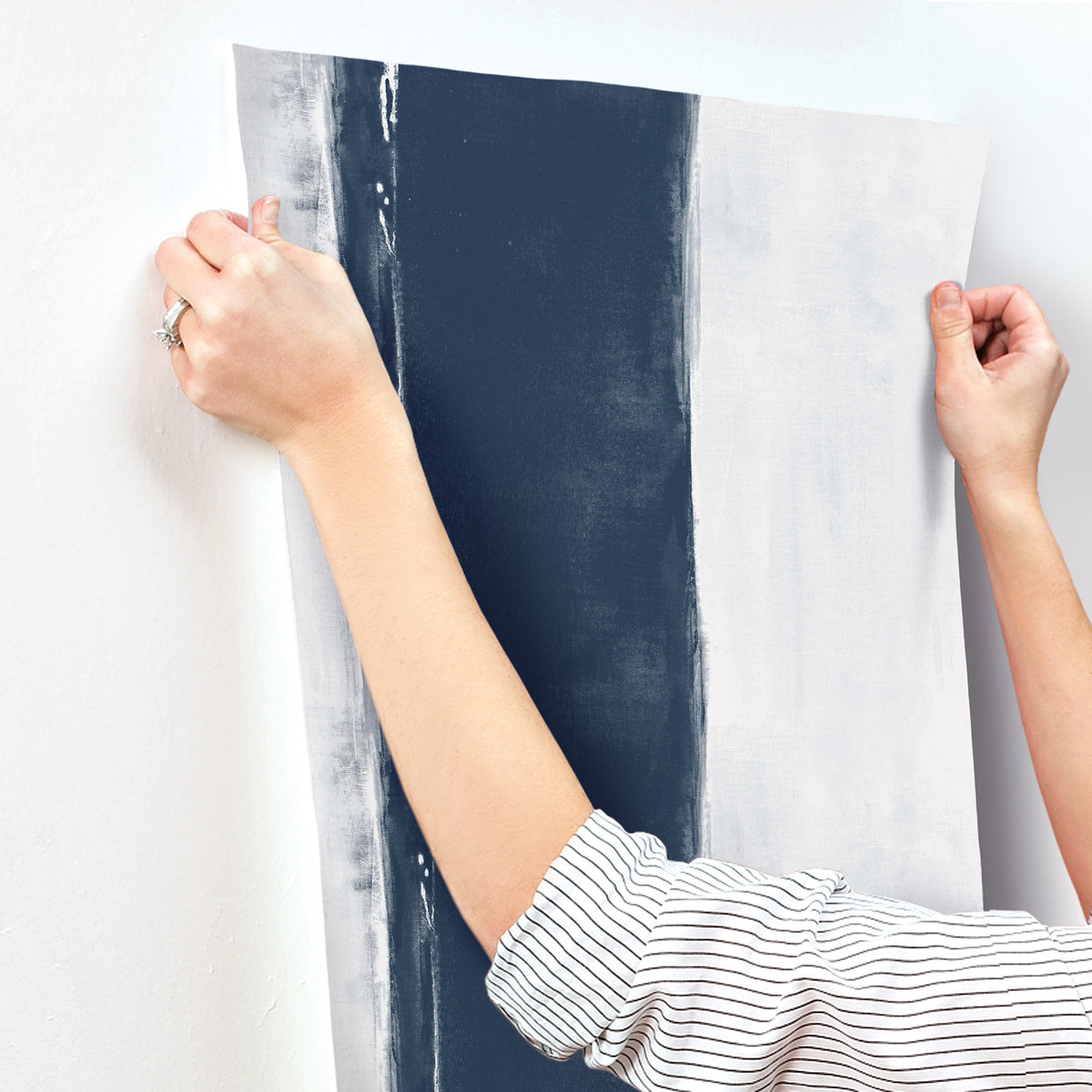 Hands holding a strip of Ink Wash Wallpaper by York Wallcoverings, featuring a wide navy blue vertical stripe on a light gray background.