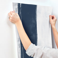 Hands holding a strip of Ink Wash Wallpaper by York Wallcoverings, featuring a wide navy blue vertical stripe on a light gray background.