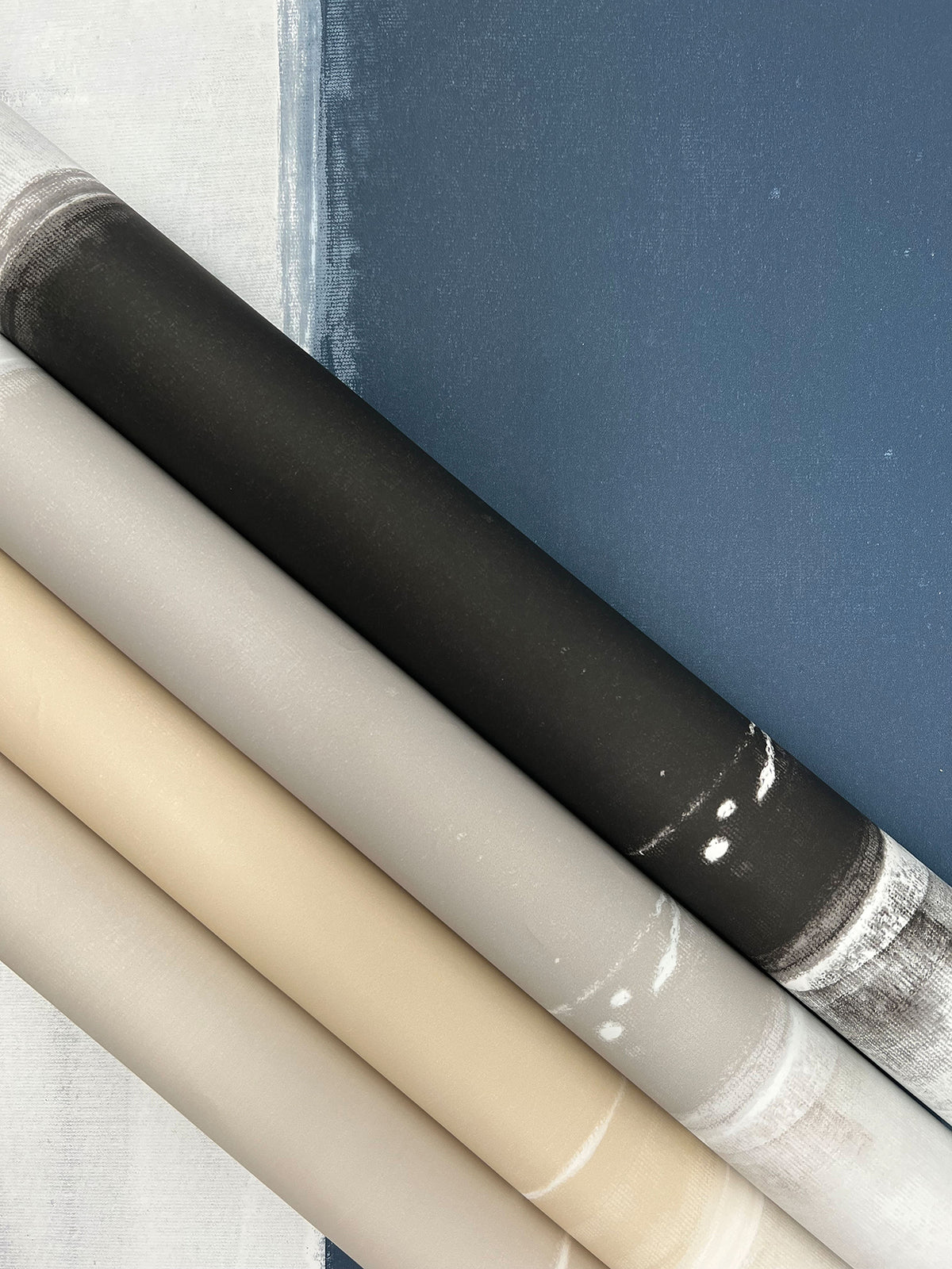 Four rolls of Ink Wash Wallpaper in shades of black, grey, beige, and light grey, layered over a blue and white painted background.