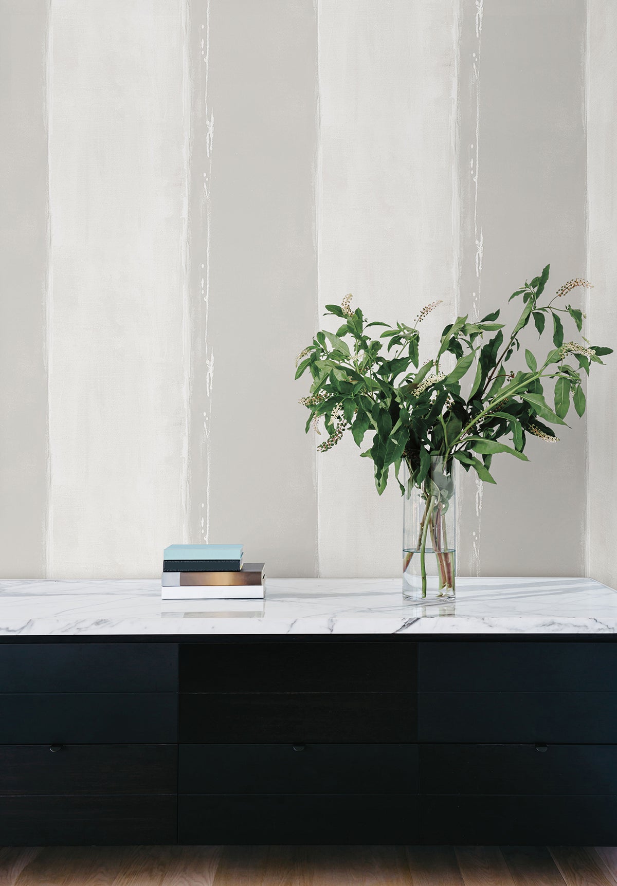 Ink Wash Wallpaper by York Wallcoverings features vertical stripes in shades of gray and white, positioned behind a black sideboard holding a glass vase of greenery and a stack of books.