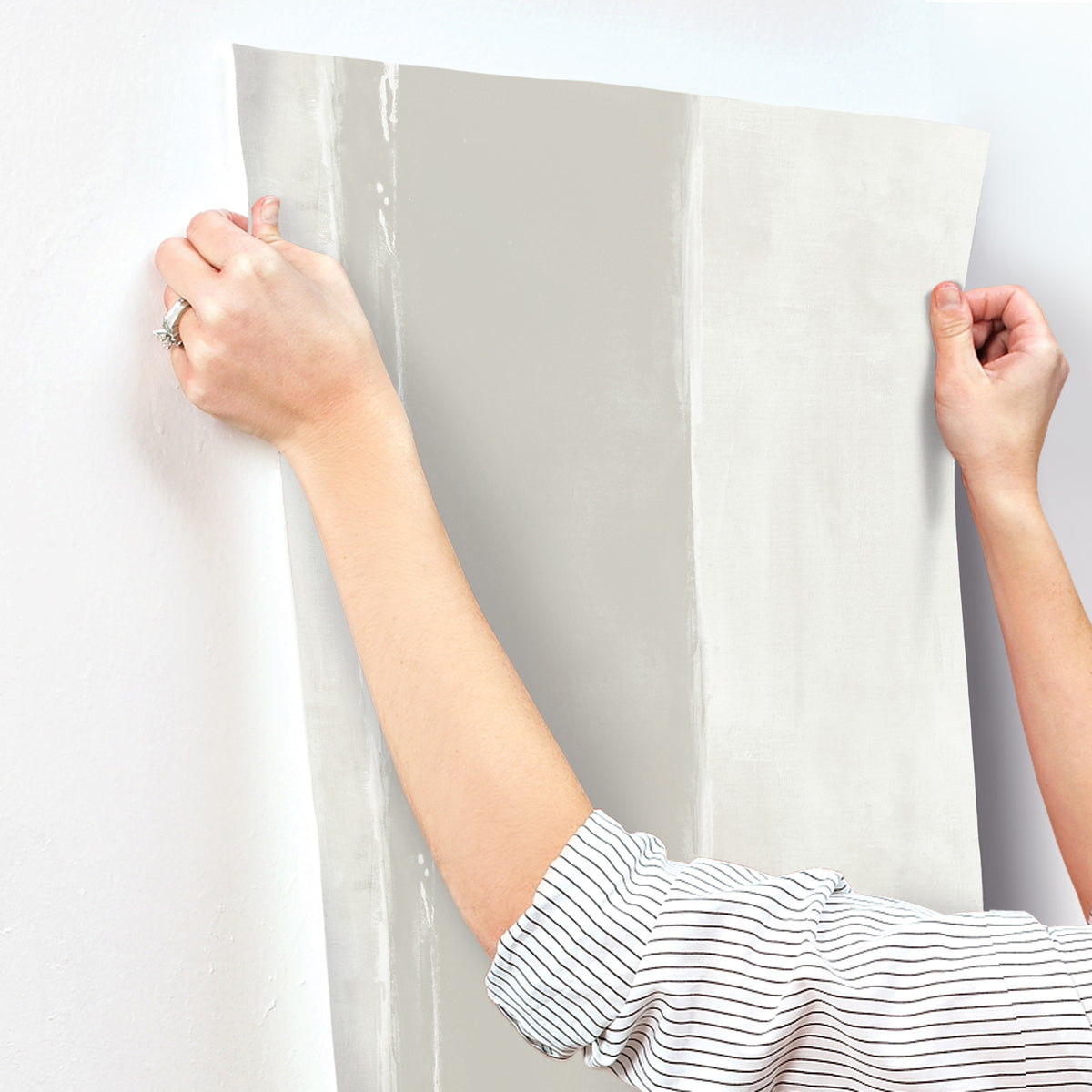 Hands holding a sheet of light gray Ink Wash Wallpaper with a vertical brushstroke pattern against a white wall.