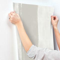 Hands holding a sheet of light gray Ink Wash Wallpaper with a vertical brushstroke pattern against a white wall.