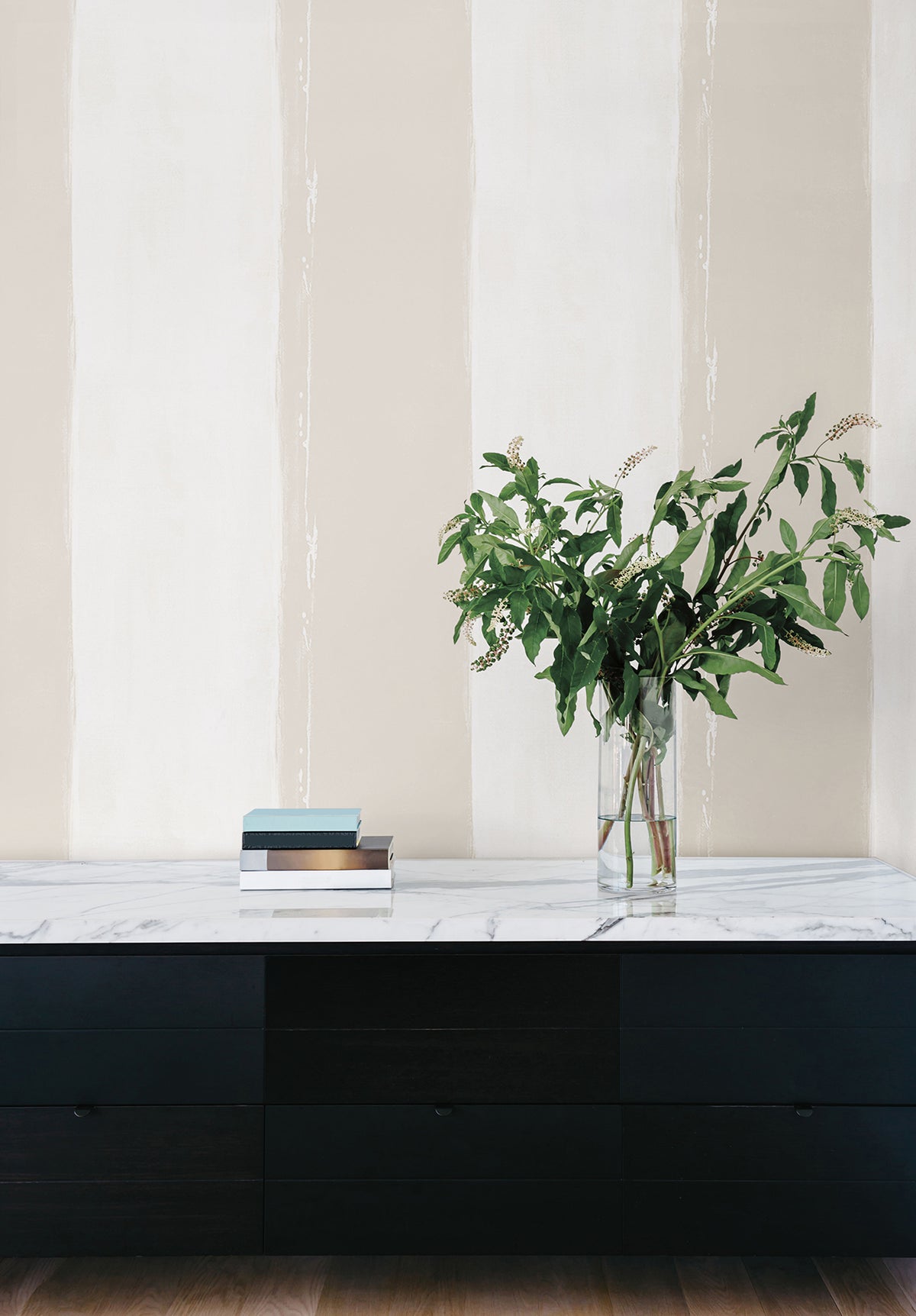 Ink Wash Wallpaper features wide, vertical brushstroke stripes in alternating shades of beige and off-white behind a black console table with a marble top, a stack of books, and a glass vase of greenery.