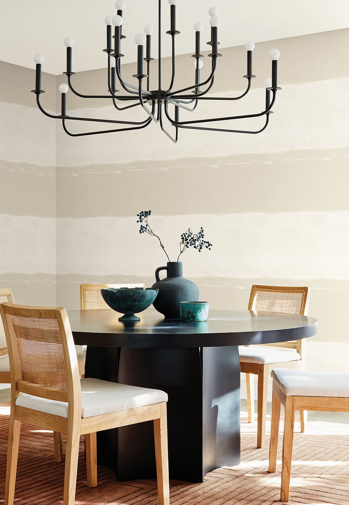 A dining room with light-colored, wide-striped Ink Wash Wallpaper by York Wallcoverings features a round black table, cane chairs, and a large black chandelier.