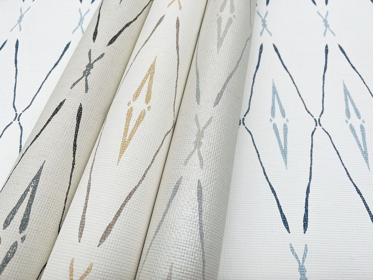 Three rolls of York Wallcoverings Artifact Silhouettes Wallpaper stand upright next to a flat sample, all featuring a repeating diamond-shaped ikat pattern in shades of blue, grey, and tan on a white textured background.