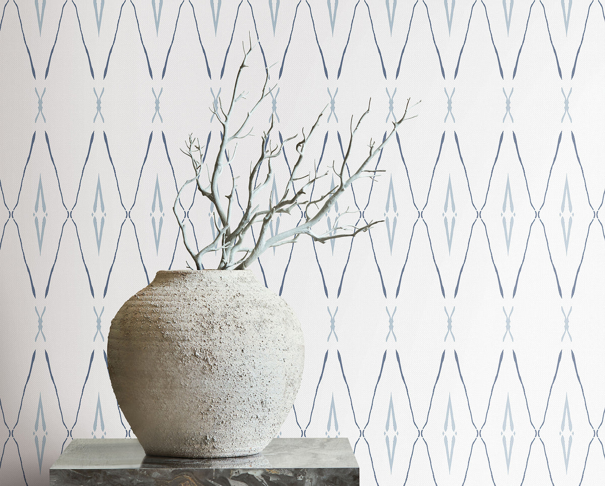 A textured stone vase with bare branches sits in front of Artifact Silhouettes Wallpaper, which features a repeating blue diamond and chevron pattern.