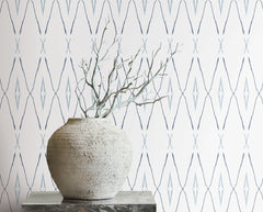 A textured stone vase with bare branches sits in front of Artifact Silhouettes Wallpaper, which features a repeating blue diamond and chevron pattern.