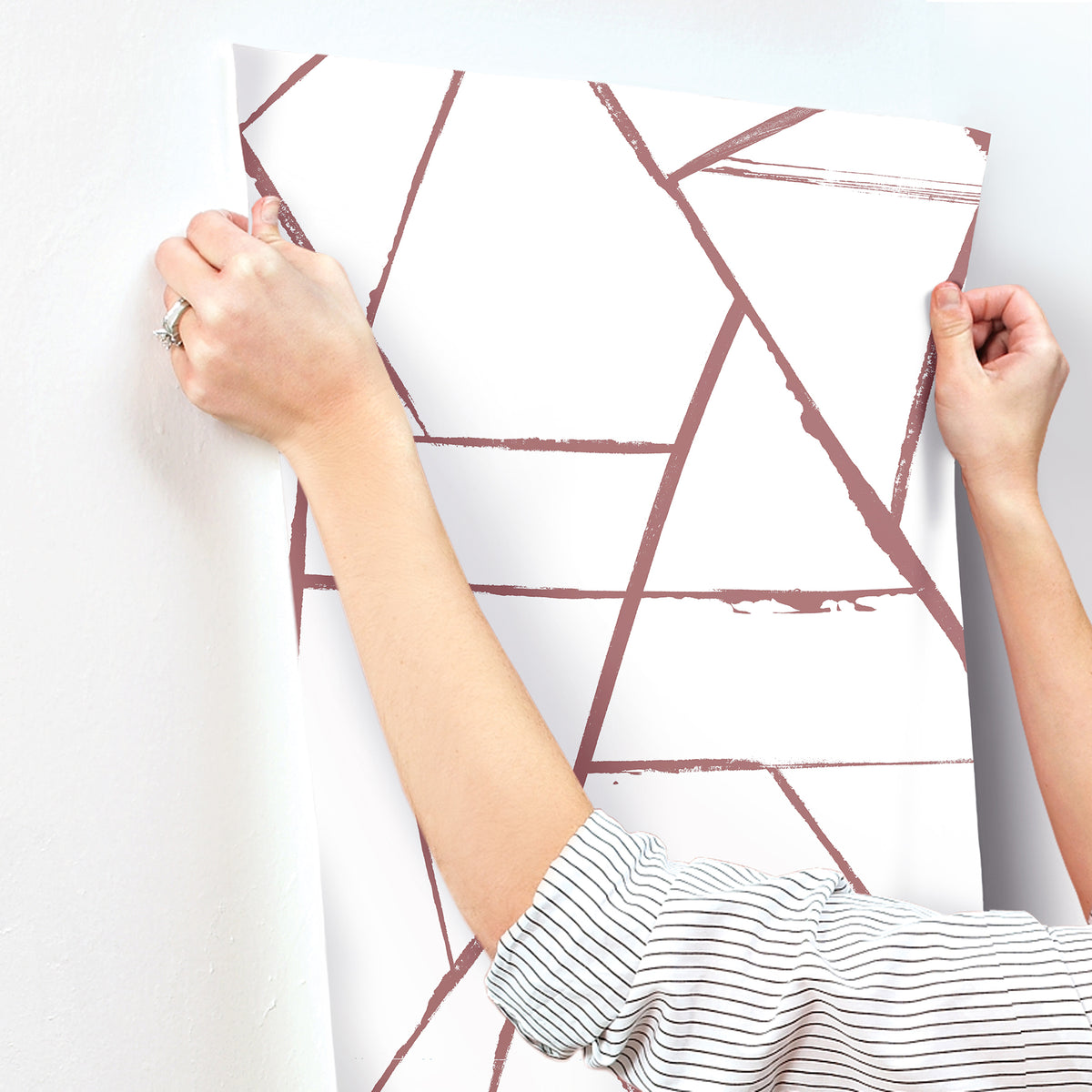 Hands applying a sheet of York Wallcoverings Intersect Wallpaper with a geometric pattern of muted pink intersecting lines to a wall.