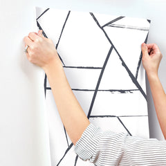 Hands applying a sheet of Intersect Wallpaper, featuring a white background with a black geometric pattern of intersecting lines, to a wall.