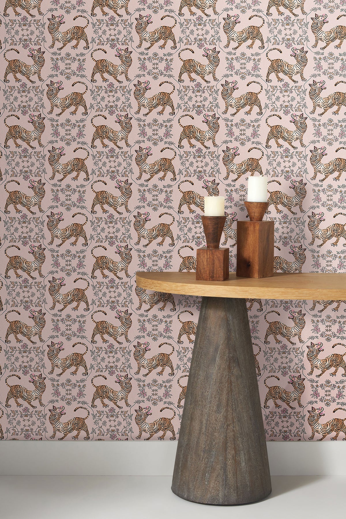 Ferrandi Wallpaper featuring a repeating pattern of tigers and floral motifs in orange, black, and white on a light pink background, next to a wooden table with two pillar candles.