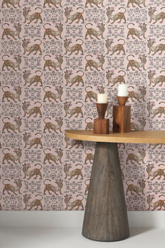Ferrandi Wallpaper featuring a repeating pattern of tigers and floral motifs in orange, black, and white on a light pink background, next to a wooden table with two pillar candles.