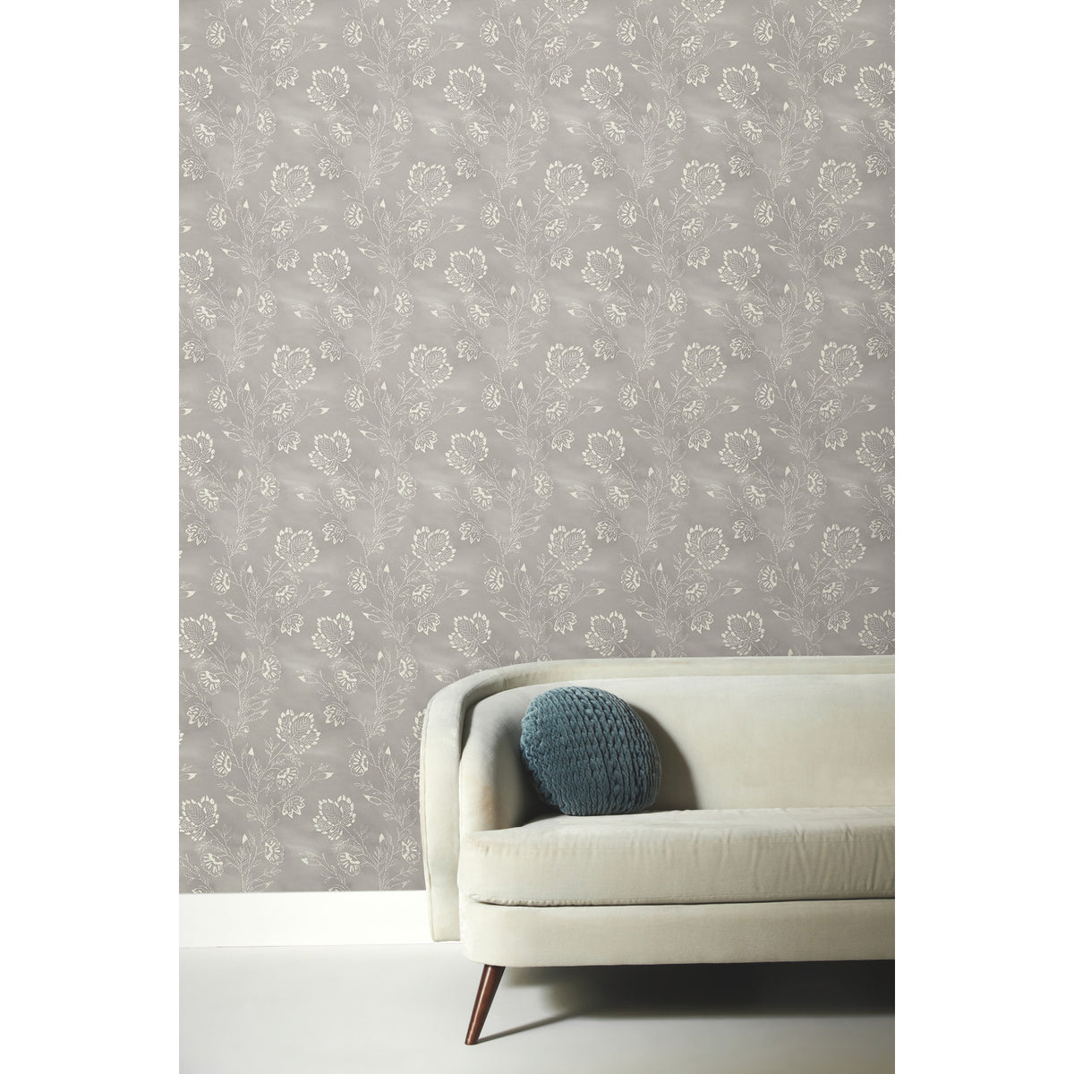 Barbier Wallpaper featuring a white botanical floral pattern on a gray background, shown behind a modern sofa with a blue round pillow.