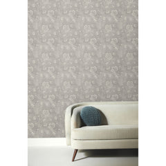 Barbier Wallpaper featuring a white botanical floral pattern on a gray background, shown behind a modern sofa with a blue round pillow.