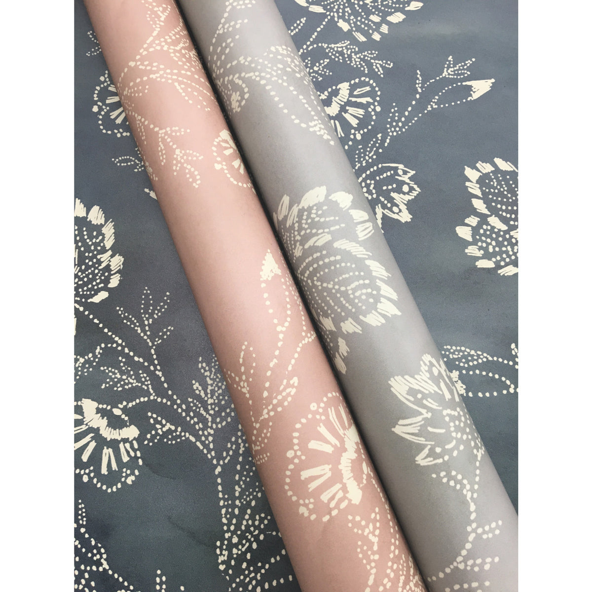 Two rolls of Barbier Wallpaper in blush pink and grey featuring a dotted floral pattern, resting on a dark grey surface with the same pattern.